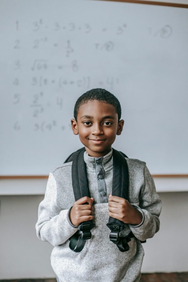 Cheerful African American boy in casual outfit with backpack standing against whiteboard in classroom and looking at camera