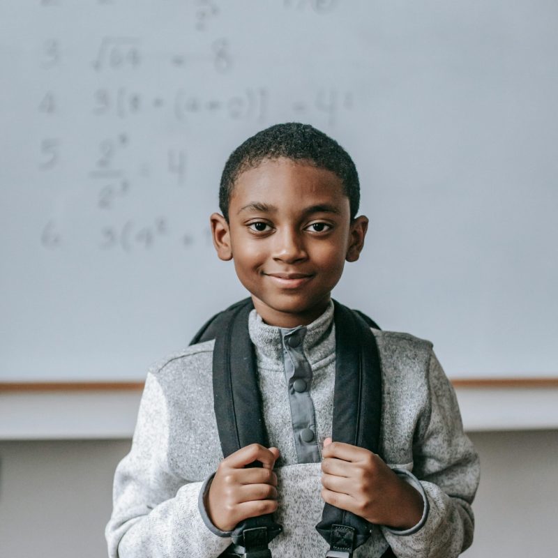 Cheerful African American boy in casual outfit with backpack standing against whiteboard in classroom and looking at camera